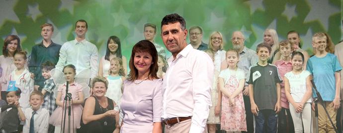 Ein großes Ensemble der Chorus-Akademie aus Kindern, Jugendlichen und Erwachsenen steht gemeinsam lächelnd auf einer Bühne mit grünem, sternenbesetztem Hintergrund. Im Vordergrund stehen die Leiter und Gründer der Chorus-Akademie Frau Tatiana Bolandeou und Herr Anatoli Fedorov.